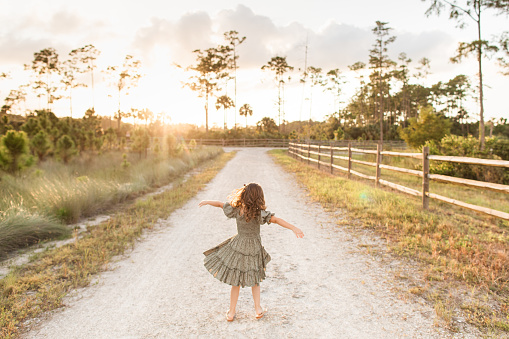 A Cute Cuban-American Toddler Girl with Brown  Curly Hair and Brown Eyes, Wearing a Green Smocked Dress, Gold Sandals & a Hair Bow, Twirling and Dancing in Nature at Sunset During Golden Hour in an Open Field with a Wooden Fence in Jupiter Farms, Florida