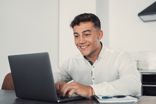 Side view handsome young businessman in eyewear working with computer remotely, sitting at wooden table in office. Pleasant happy man communicating in social network, searching information online.