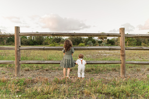 Cute Cuban-American Brother and Sister with Brown  Hair and Brown Eyes Walking, Discovering Nature at Sunset in an Open Field with a Wooden Fence in Jupiter Farms, Florida