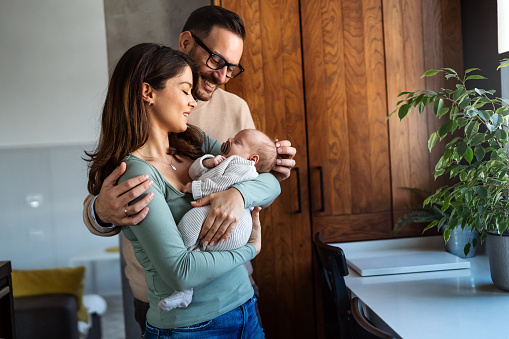 Portrait of young happy man and woman holding newborn cute babe dressed in white unisex clothing. Caucasian smiling father and mother embracing tenderly adorable new born child. Happy family concept