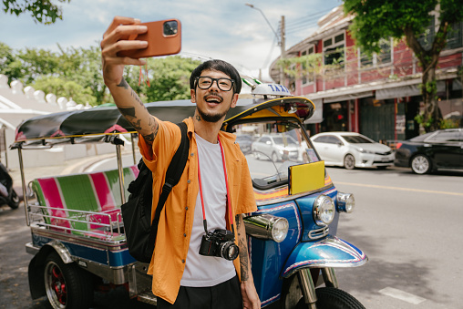 Young Asian man enjoying traveling and taking selfies with his smartphone. The background is a Tuk-Tuk taxi of Thailand.