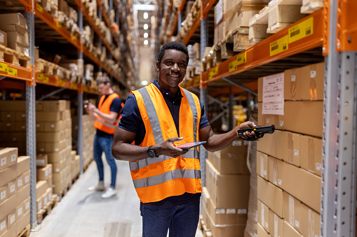 Smiling african man wearing reflective clothing working at distribution warehouse. Happy male warehouse worker looking at camera while scanning boxes on rack.
