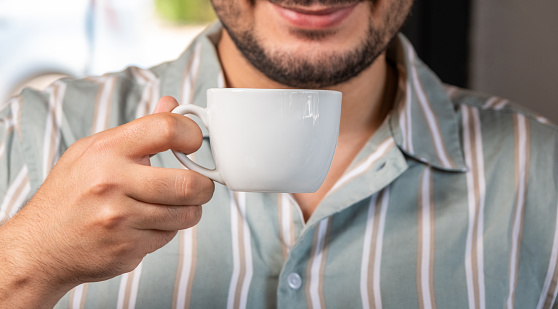 A man drinks coffee while smiling. Close-up. White coffee cup. The man is wearing a striped shirt. The man's face is not visible.