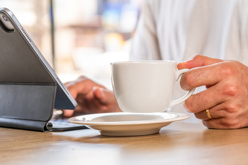 Man drinking coffee while working. Man drinks coffee in cup while using tablet computer. He consumes hot drinks. Close-up. White T-shirt. Pleasant business world.