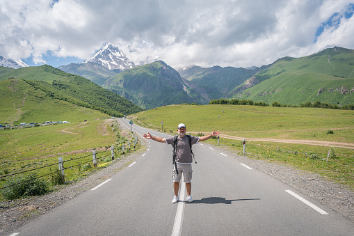 Happy traveler man opening hands while walking on mountain road away from camera