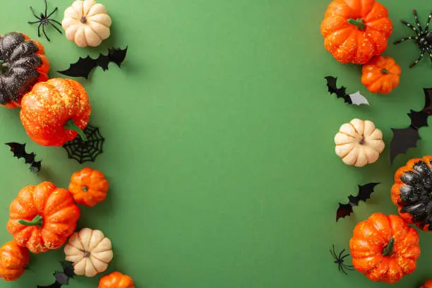 Photo of Original Halloween theme. Top-down view of themed embellishments, petite pumpkins, insects, eerie spiders, spiderweb, bats on green surface, vacant space for text message or promotion