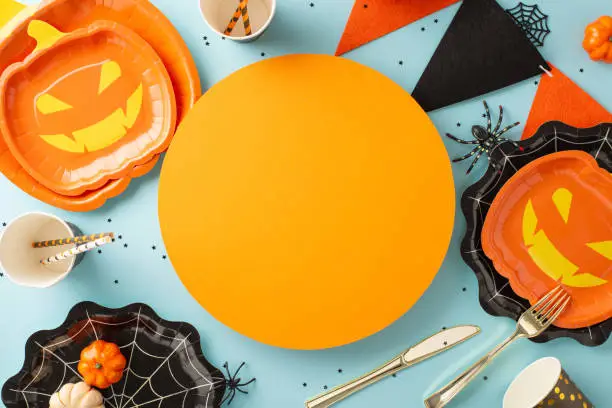Innovative Halloween feast layout. Top view of imaginative pumpkin & spiderweb plates, utensils, disposable cups, straws, pumpkins, eerie spider, garland, confetti on soft blue with circle for text