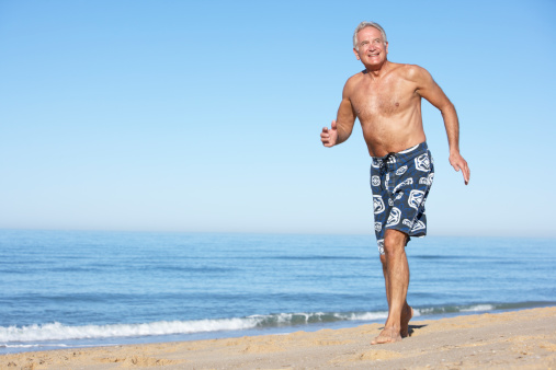 Senior Man Running Along Summer Beach Wearing Swim Shorts