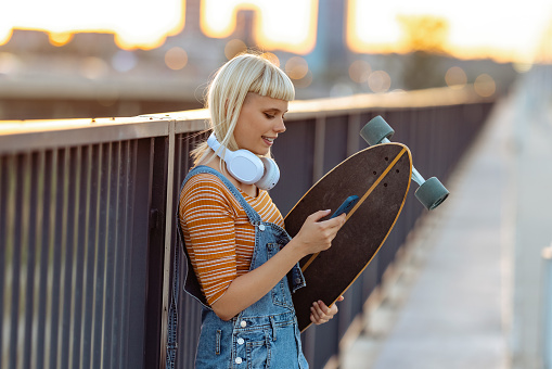 Side shot of a young woman with blond hair holding a longboard and using smart phone on the street.