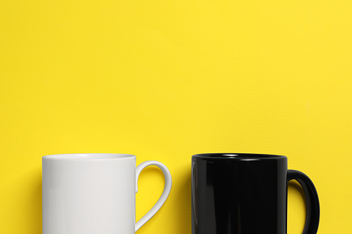 Black and white ceramic mugs on yellow background, flat lay. Space for text