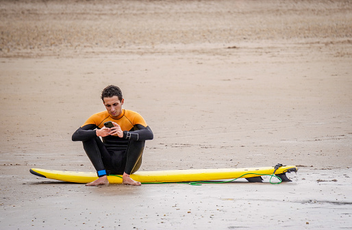 Tired Injured Surfer with Upset Face Sitting on his Surfboard on the Ground at the Beach and Looking at his Phone