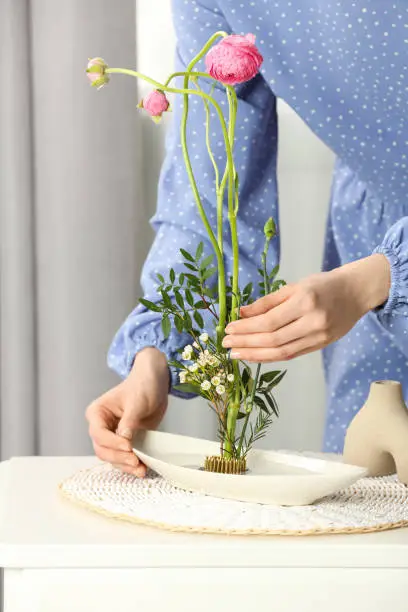 Woman creating stylish ikebana with beautiful flowers and green branches at white table, closeup