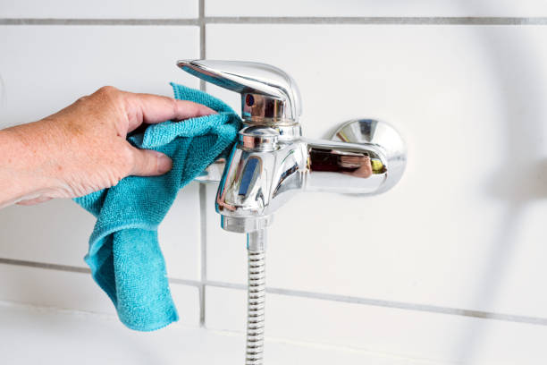 Cleaning a bathtub faucet with a blue microfiber cloth to remove limescale stains from hard calcium water, copy space, selected focus, narrow depth of field stock photo