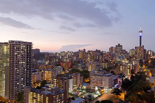 Aerial view of the city of Johannesburg at sunset, with the tallest building being the Telkom Tower which has its lights on at the top.  Most of the buildings are of modern design.  The sun is setting to the left of the city.