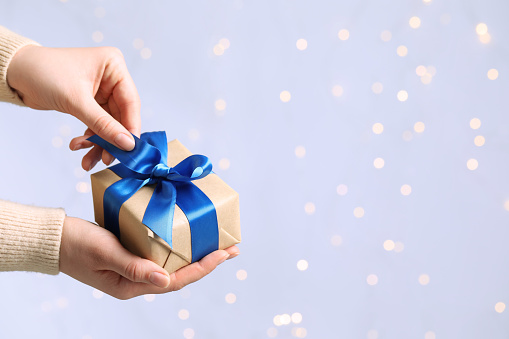 Woman holding gift box with blue bow against blurred festive lights, closeup and space for text. Bokeh effect