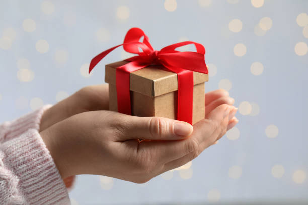 istock Woman holding gift box with red bow against blurred festive lights, closeup. Bokeh effect 1664209028