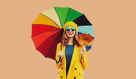 Autumn color style outfit, beautiful smiling young woman holds yellow maple leaves with colorful umbrella posing in french beret hat, coat on beige studio background