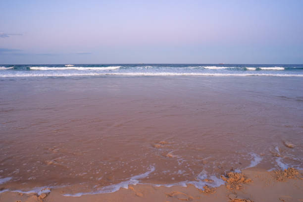 The foreshore covered by water as the tide rises on a sandy surf beach showing surf, waves, breaking water,and ships on the distant horizon at dusk