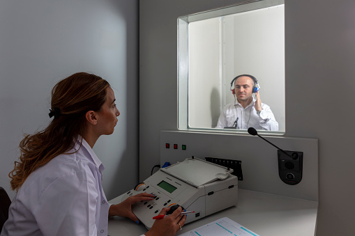 A female audiologist conducting a hearing examination on a male patient using an audiometer in a private sound room. Test room in hospital.