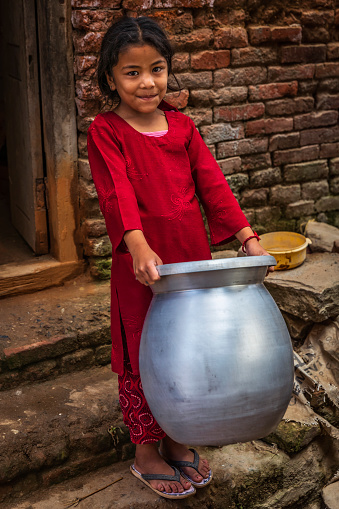 Portrait of young Nepali girl crrying a big metal pot in an ancient town of Bhaktapur. Bhaktapur is an ancient town in the Kathmandu Valley and is listed as a World Heritage Site by UNESCO for its rich culture, temples, and wood, metal and stone artwork.