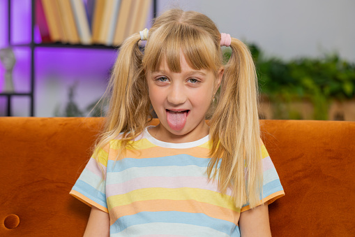 Caucasian joyful preteen school girl making playful silly facial expressions and grimacing, fooling around, showing tongue. Young excited blonde child kid at home room apartment sitting on couch