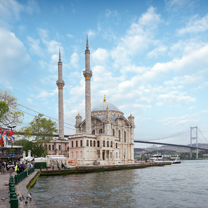 Istanbul, Turkey - May 12, 2023: Ortakoy Mosque at midday, bathed in bright sunlight, with minarets and domes glistening in the rays, and Bosphorus Bridge visible in the background