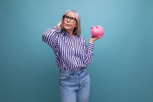 smart 60s woman with gray hair holding piggy bank on bright studio background.