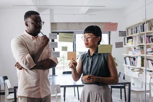 Two pensive entrepreneurs with glasses (Japanese female and African-American male) discussing ideas and brainstorming while standing in the office.