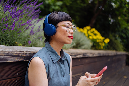A wide angle view of a smiling Japanese female with glasses (eyes closed) using an app on her smartphone and her wireless headphones while relaxing in the park.