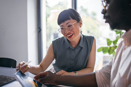 A beautiful Japanese woman with glasses explaining some statistics to her African-American coworker while they are sitting at the desk. (mentorship concept)