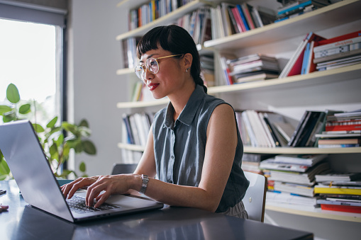 A wide angle view of a smiling Japanese entrepreneur typing on her laptop while sitting at the office desk.