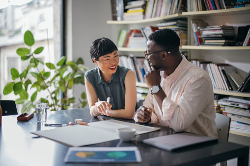 A laughing Japanese businesswoman having a conversation with her African-American coworker while they are sitting at the desk.