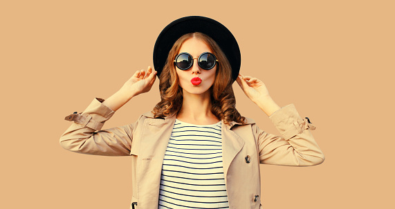 Portrait of beautiful young woman posing blowing her lips sends sweet air kiss wearing sunglasses, black round hat on beige studio background