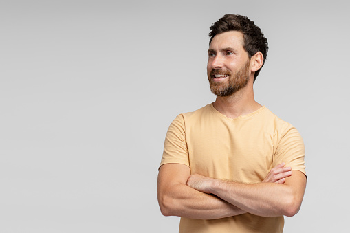 Smiling handsome bearded man, hipster wearing blank t shirt looking away and posing arms crossed isolated on grey background. Happy fashion model posing for pictures, studio shot. Mockup