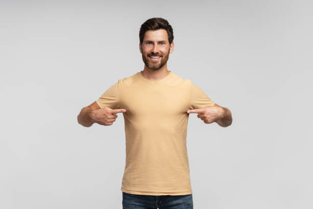 istock Smiling bearded man wearing blank t shirt pointing himself isolated on gray background. Mockup 1664568096