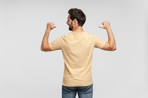 Back view of handsome bearded man, hipster wearing blank t shirt pointing himself isolated on gray background. Happy successful fashion model posing for pictures, studio shot. Mockup