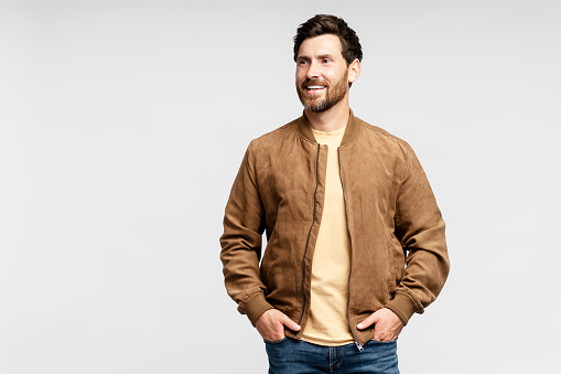 Handsome smiling bearded man wearing brown autumn jacket, stylish jeans isolated on grey background. Portrait of successful middle aged fashion model posing for pictures, studio shot