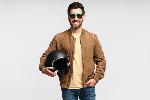 Handsome smiling bearded man, biker holding motorcycle helmet isolated on gray background. Fashion model wearing brown leather jacket, stylish sunglasses looking at camera, posing for pictures