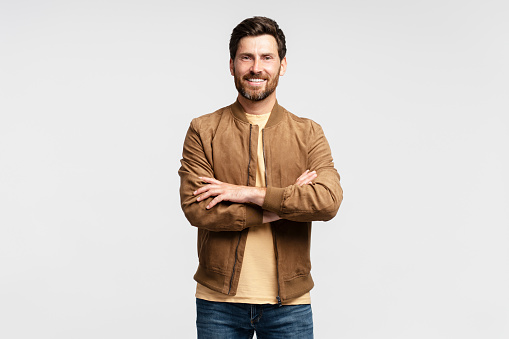 Handsome smiling bearded man wearing brown autumn jacket looking at camera isolated on grey background. Portrait of successful middle aged fashion model posing for pictures, studio shot