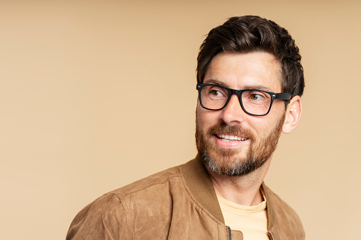 Portrait of smiling handsome bearded man wearing stylish eyeglasses looking away isolated on beige background, copy space. Vision concept