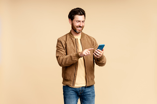Portrait of smiling bearded man holding mobile phone, reading text message, communication online isolated on beige background. Handsome guy shopping online, choosing on website