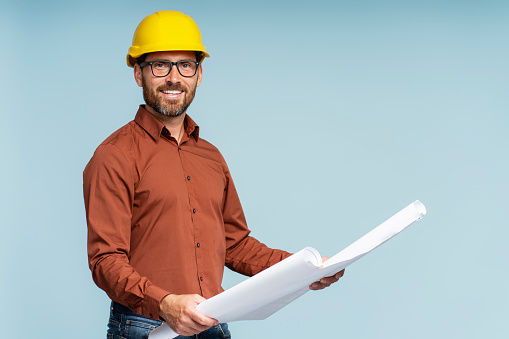 Portrait of handsome smiling architect or engineer wearing yellow hardhat holding architectural drawing looking at camera isolated on blue background. Successful business, career concept
