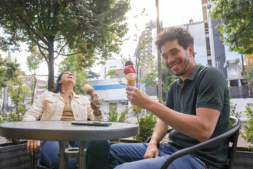 A latin couple shares ice cream while sitting near the street. The man looks at the camera while the girl smiles spontaneously.