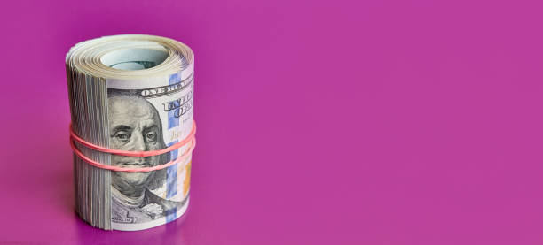 istock Many hundred dollar bills rolled up with a red rubber band on a colored background. 1664924525