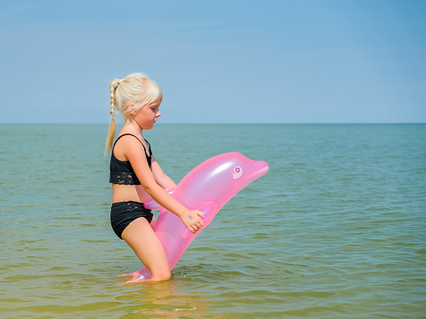 Little cute blonde baby girl swims in the sea with an inflatable pink dolphin. A child plays and swims in the sea on a sunny summer day. Happy child is resting on the sea.