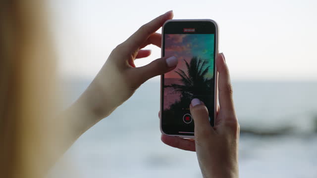 Woman holds smartphone and recording nature of tropical island. Phone screen with video of blue rocky ocean, blue cloudy sky and green palm trees landscape. Blogger enjoys filming views on holidays.