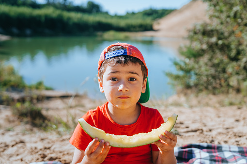 Sad caucasian little boy in casual finished eat watermelon sitting on plaid against lake on summer sunny day, give me more. Tourism, family leisure, childhood. Cute American kid at outdoor trip.