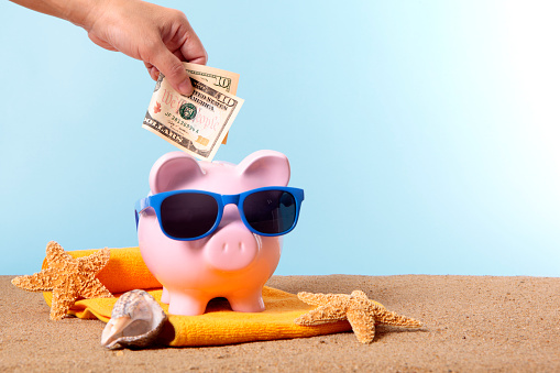 Saving for beach vacation, with pink piggy bank and sunglasses.  Studio shot with plain blue background.  Sharp focus on the ten dollar bill.  Space for copy.  Warm color and directional lighting are intentional.