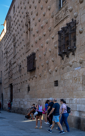 Street violinist playing music and tourists walking along the street of La Casa de las Conchas facade with wrought iron windows in Salamanca in Spain.