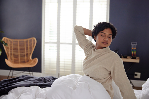 A young woman sits up in bed, stretching, and the setting could be either at home or in a hotel room.